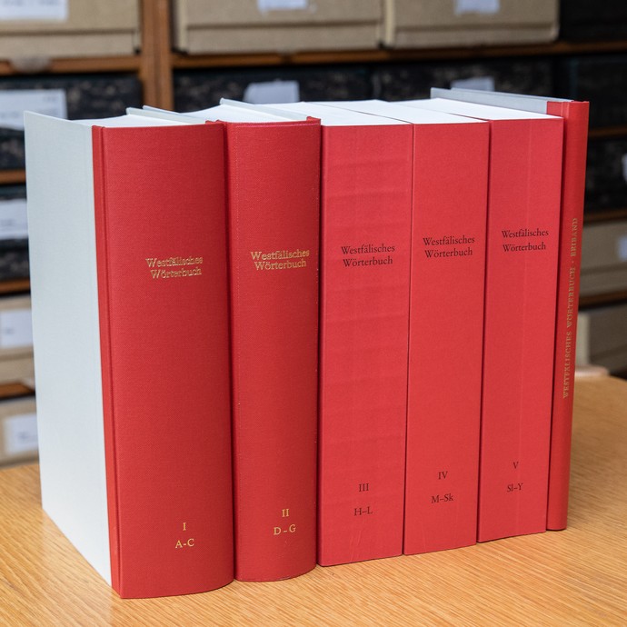 alle Bände des Westfälischen Wörterbuchs auf einem Tisch, im Hintergrund die Zettelkästen des Wörterbucharchivs (öffnet vergrößerte Bildansicht)