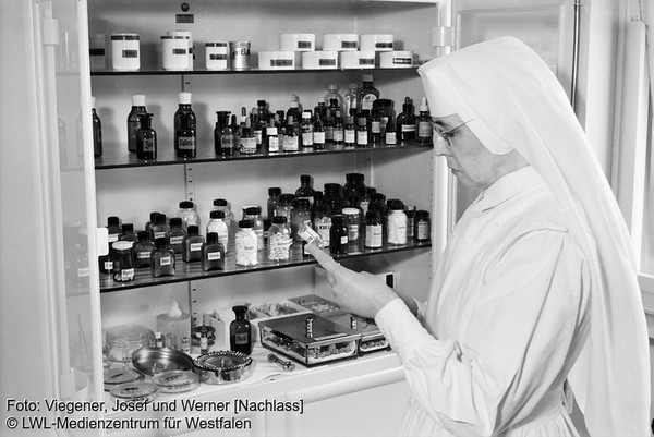Als Schwarz-Weiß-Foto ist eine Frau, vermutlich eine Apothekerin, im weißen Kittel dargestellt, wie sie vor einem vollen Medizinschrank steht.