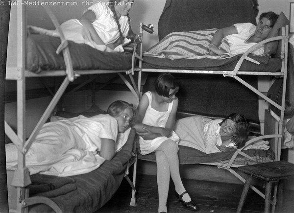 Mehrere Frauen in Schlafkleidung unterhalten sich von zwei Etagenbetten aus, scheinbar während einer Übernachtung in einer Jugendherberge.